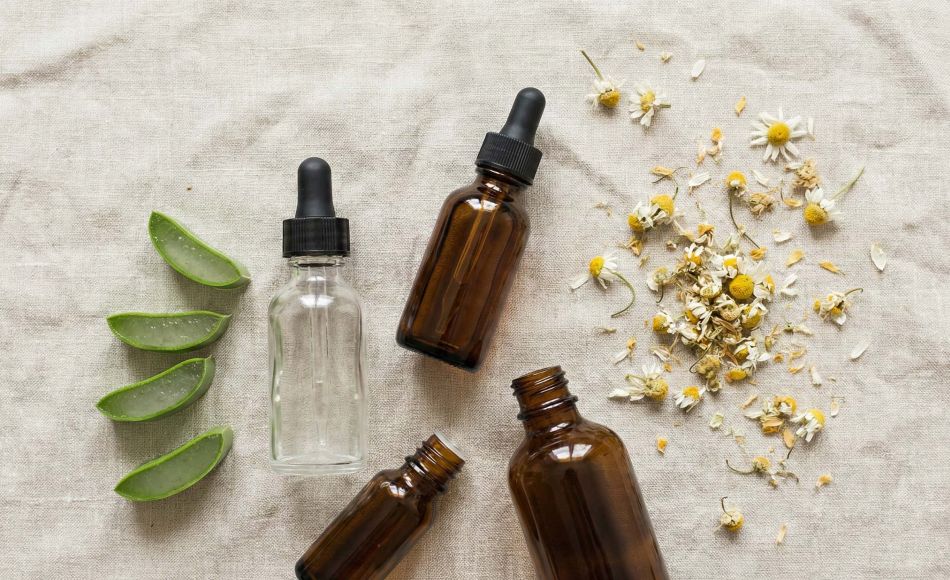 A collection of natural skincare ingredients including aloe vera, chamomile, and botanical oils on a light pink linen surface.