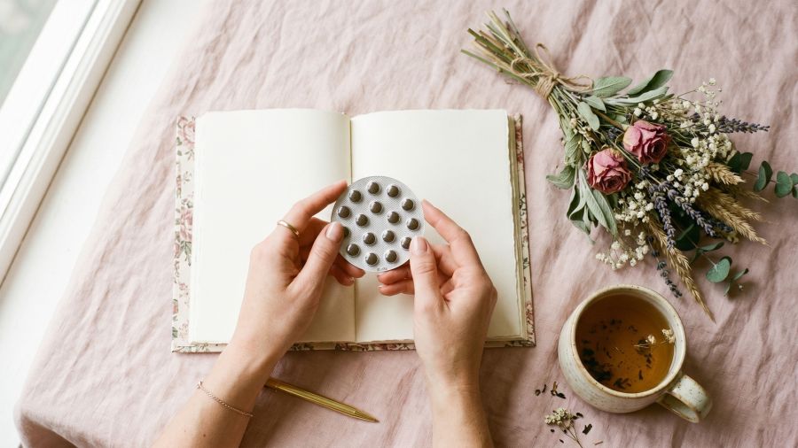 Woman's hands holding birth control pill pack beside a journal and herbal tea on a pastel background