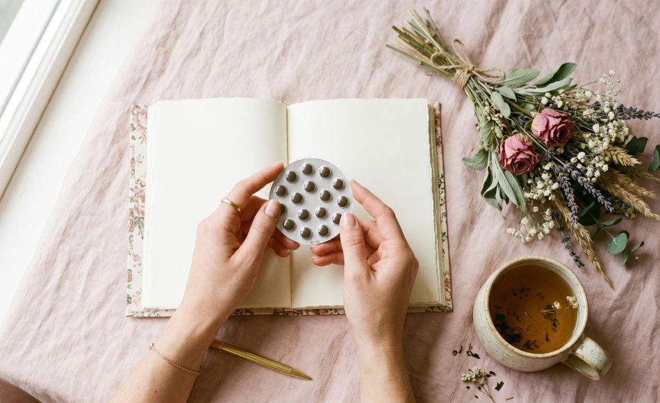 Woman's hands holding birth control pill pack beside a journal and herbal tea on a pastel background