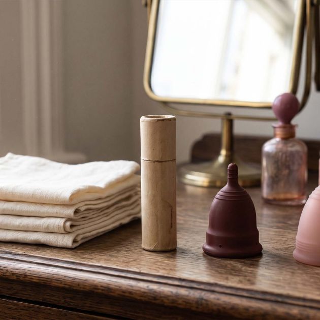 History of period products timeline shown on a sunlit dressing table with folded linen cloths, a vintage cardboard applicator, a rubber menstrual cup and a modern silicone cup