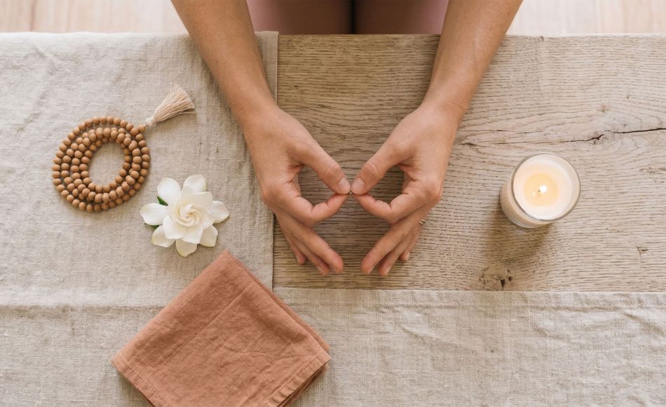 Woman's hands resting in a meditation mudra beside mala beads, a white flower, and a candle on a linen surface