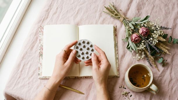Woman's hands holding birth control pill pack beside a journal and herbal tea on a pastel background