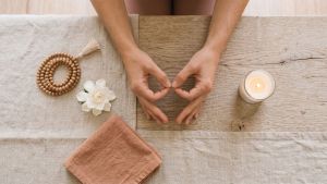 Woman's hands resting in a meditation mudra beside mala beads, a white flower, and a candle on a linen surface