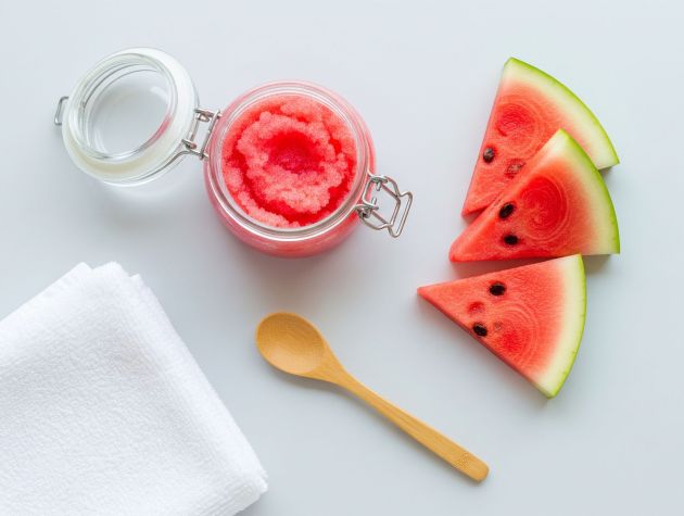 Jar of pink watermelon sugar scrub with fresh watermelon slices and wooden spoon on a pale background.